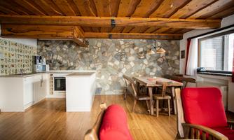A cozy living kitchen with wooden ceilings and a stone wall. There is a dining area with wooden tables and chairs, as well as red relaxation chairs.
