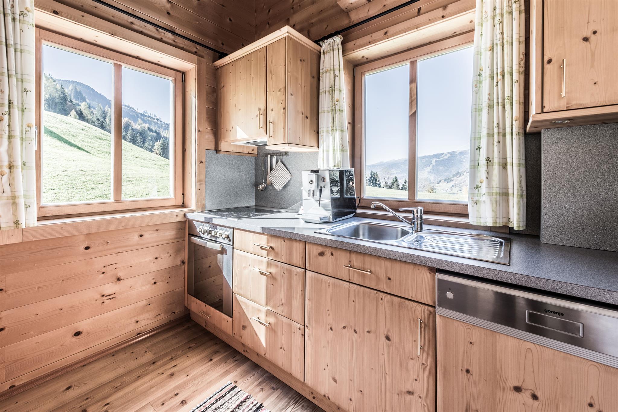 A modern kitchen made of wood with large windows and a view of nature. The surroundings are designed to be bright and friendly.