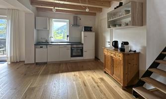 A modern kitchen with wooden floors and light-colored walls. In the background, there are windows and a staircase visible.
