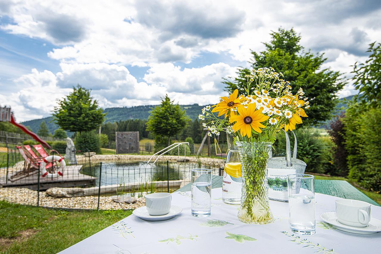 A beautiful garden with a small pool and green trees. On a table, there is a vase with sunflowers and glasses.