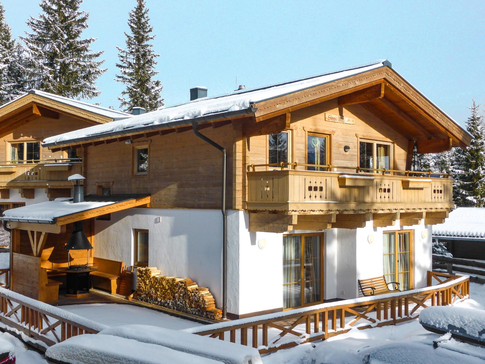 A cozy wooden house in the snow, surrounded by tall fir trees. The balcony and the veranda invite you to relax.