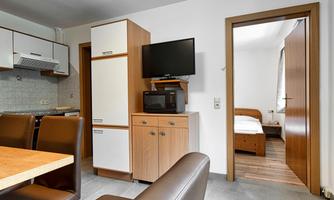 A modern kitchen with a dining table and a television. In the background, there is a bedroom with a bed.