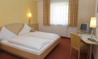 A simple room with a double bed and two pillows. There is a television and a chair at the table, with curtains that provide natural light.