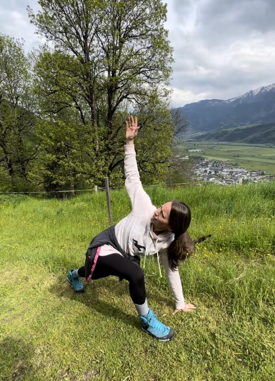 A person is doing a stretching exercise outdoors on a green meadow. In the background, trees and mountains are visible.
