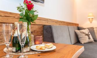 A table with two champagne glasses, bottles of sparkling wine, and a plate with small snacks. In the background, there is a vase with red roses.