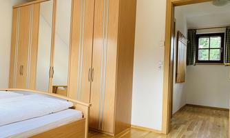 A bright bedroom with a wooden wardrobe and a bed that has white bedding. In the background, there is a hallway with a window.