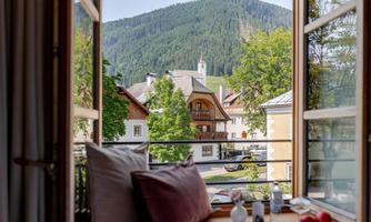 View from a window overlooking a picturesque village with mountains in the background. The surroundings are green and inviting, perfect for a relaxing stay.