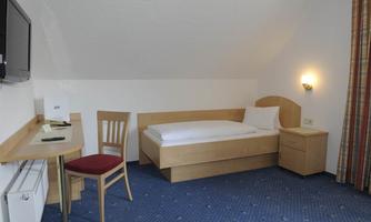 A simple single room with a bed, a table, and a chair. The room is bright and well-equipped with a television and a nightstand.