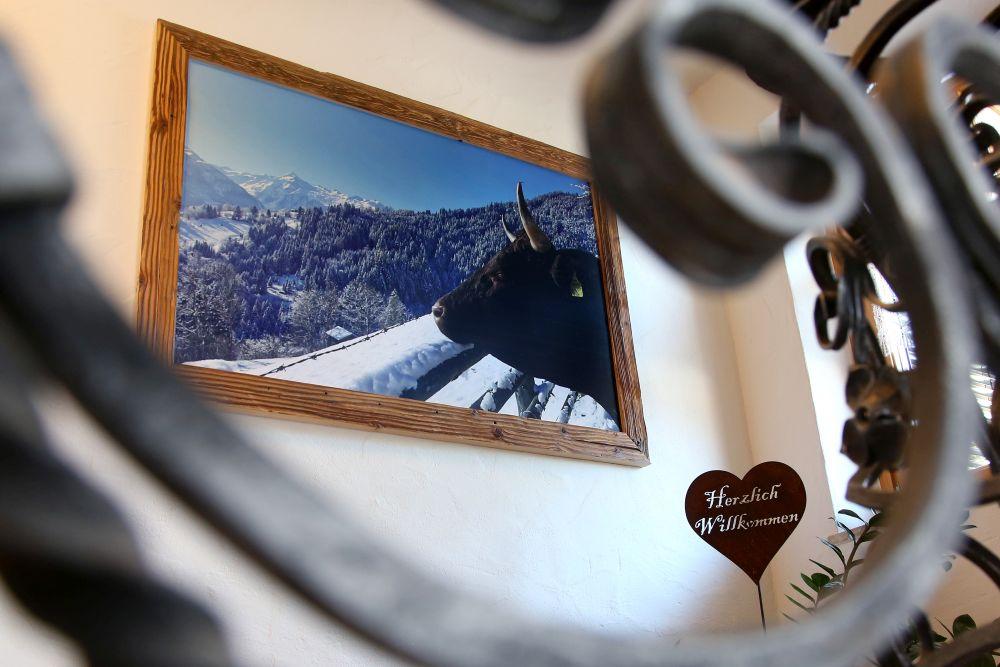 An image of a house with a magnificent view of the mountains and a cow on a windowsill. A heart-shaped sign that reads "Welcome" complements the atmosphere.