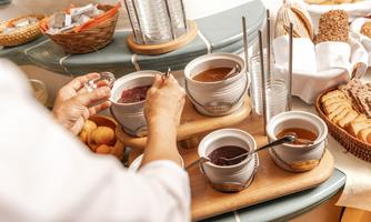 A table with various jams in bowls and a selection of fresh bread. A hand is stirring in one of the bowls.
