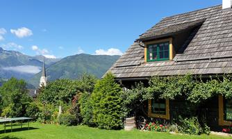 A picturesque house surrounded by a green garden and mountains in the background. The sky is clear and the landscape is idyllic.
