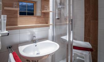 A modern bathroom with a shower and a sink. The walls are light tiled, and there are light wooden shelves as well as red towels.