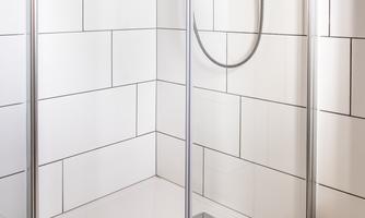 A modern shower cubicle with white tiles and wooden flooring. The shower head is mounted on the wall and the cubicle has glass walls.