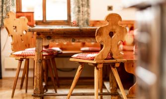 A rustic wooden dining table with colorful seat cushions. In the background, additional wooden chairs and a window can be seen.