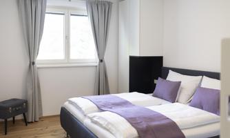 A modern bedroom with a double bed and purple decorative pillows. Large windows let in plenty of daylight, and the walls are painted in neutral colors.