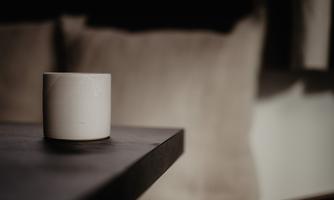 A white cup is sitting on a dark table. Soft pillows are visible in the background.