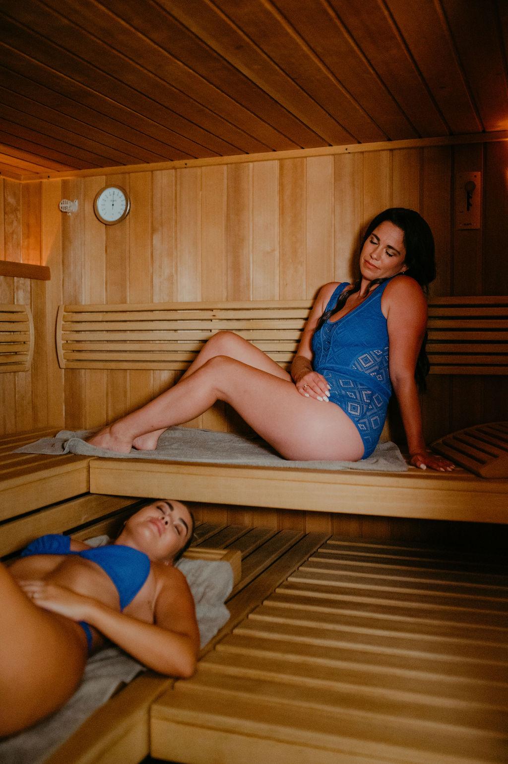 Two women are relaxing in a sauna. One is sitting on the upper bench, while the other is lying on the lower bench.