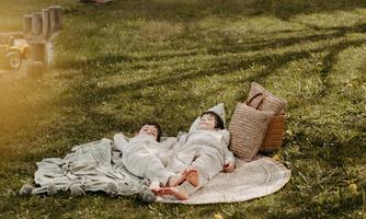 Two children are lying relaxed on a blanket in the grass. Next to them is a basket, and the sun is shining on the scene.