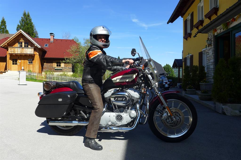A motorcyclist is sitting on a red Harley-Davidson in front of a wooden house. The scene is captured in sunny weather with a clear sky.