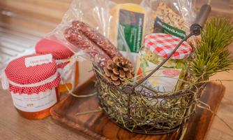 A decorative basket with various local foods, including sausage, cheese, and jars of jam. The basket is placed on a wooden board and also contains a small pine cone decoration.