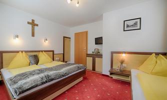 A cozy hotel room with two beds, yellow pillows, and a red carpet. There is a cross hanging on the wall, and there is a television as well as a small table.