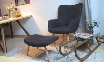 A stylish armchair with a footrest is located in a cozy room. Next to a wooden table and a rustic coffee table with flowers and magazines.