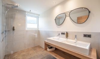 A modern bathroom with a large shower and a double sink. Above the sink hangs a striking mirror in the shape of sunglasses.