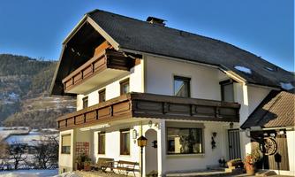 A cozy multi-story house with wooden balconies in a winter landscape. Snow-covered mountains can be seen in the background.