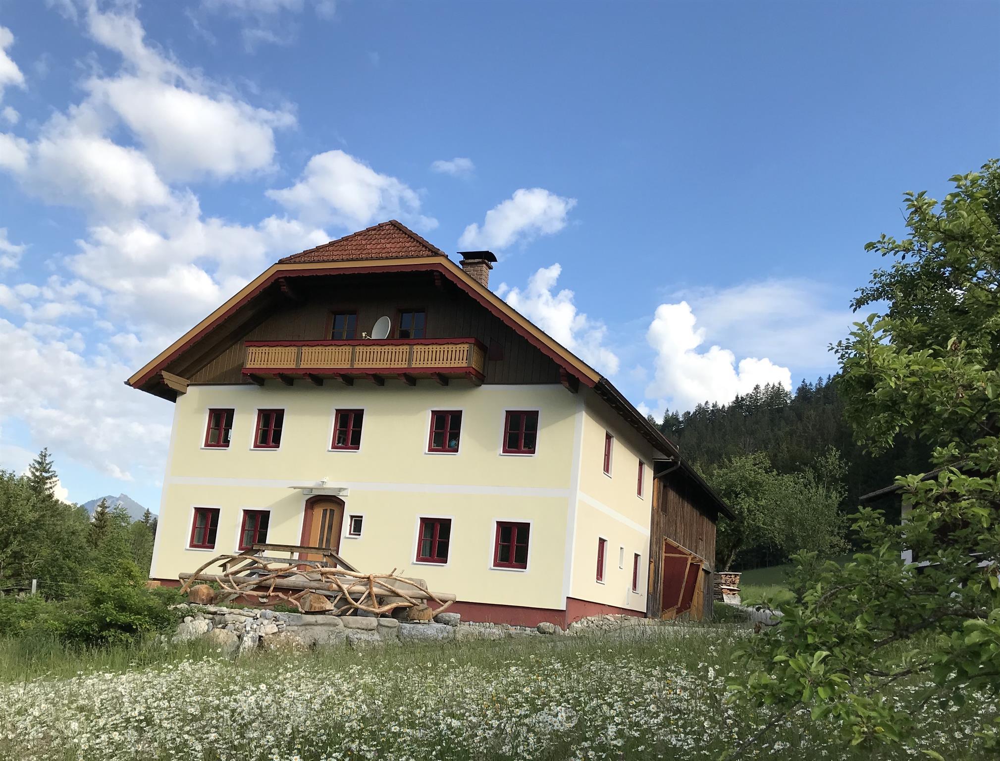 A large, modern house in nature with a wooded area in the background. The meadow is lined with flowers and the sky is partially cloudy.