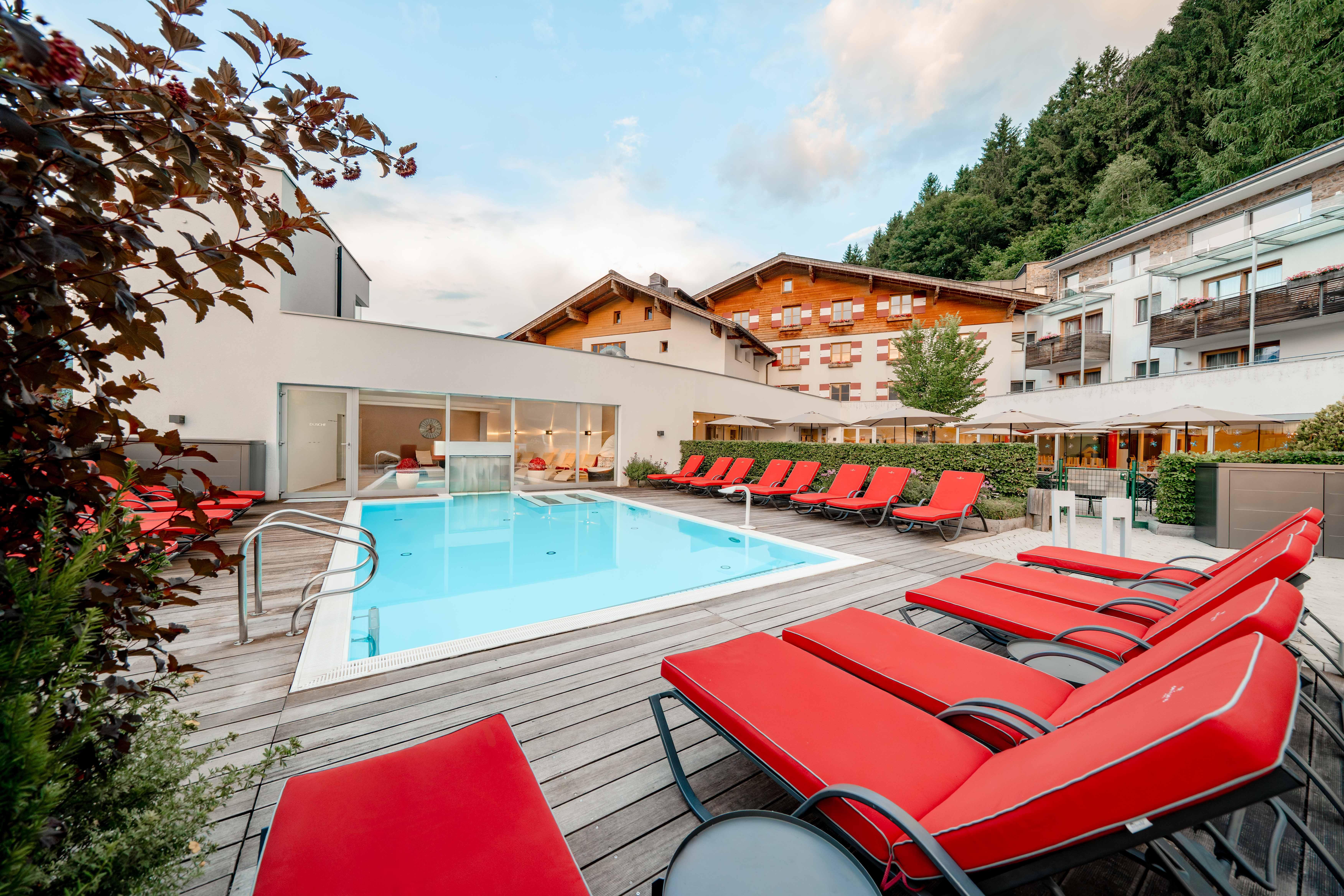 A modern pool area with red loungers and clear water. In the background, some buildings and green trees are visible.