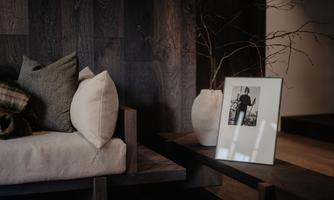 A cozy living room with a wooden sofa and various cushions. Next to the sofa, there is a vase and a framed picture on a table.