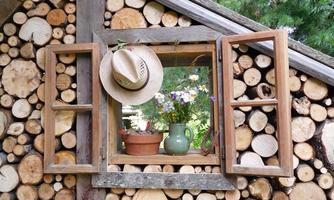 A wooden house with a window opening decorated with a pot of flowers and a green vase. A hat hangs over the window, surrounded by wooden logs.