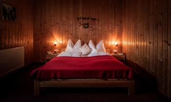A cozy bedroom with a wooden wall design. The bed is decorated with red blankets and white pillows, while soft light creates a warm atmosphere.