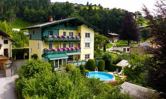 A beautiful guesthouse with colorful flowers on the balcony. In the foreground is a pool and in the background are green hills.