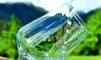 A glass is being held while water splashes over it. In the background, green meadows and mountains can be seen.