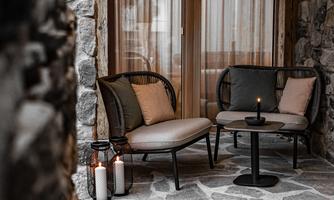A cozy seating area with a table and two chairs in a room with stone walls. In front of a window with curtains, there is a candle on the table.