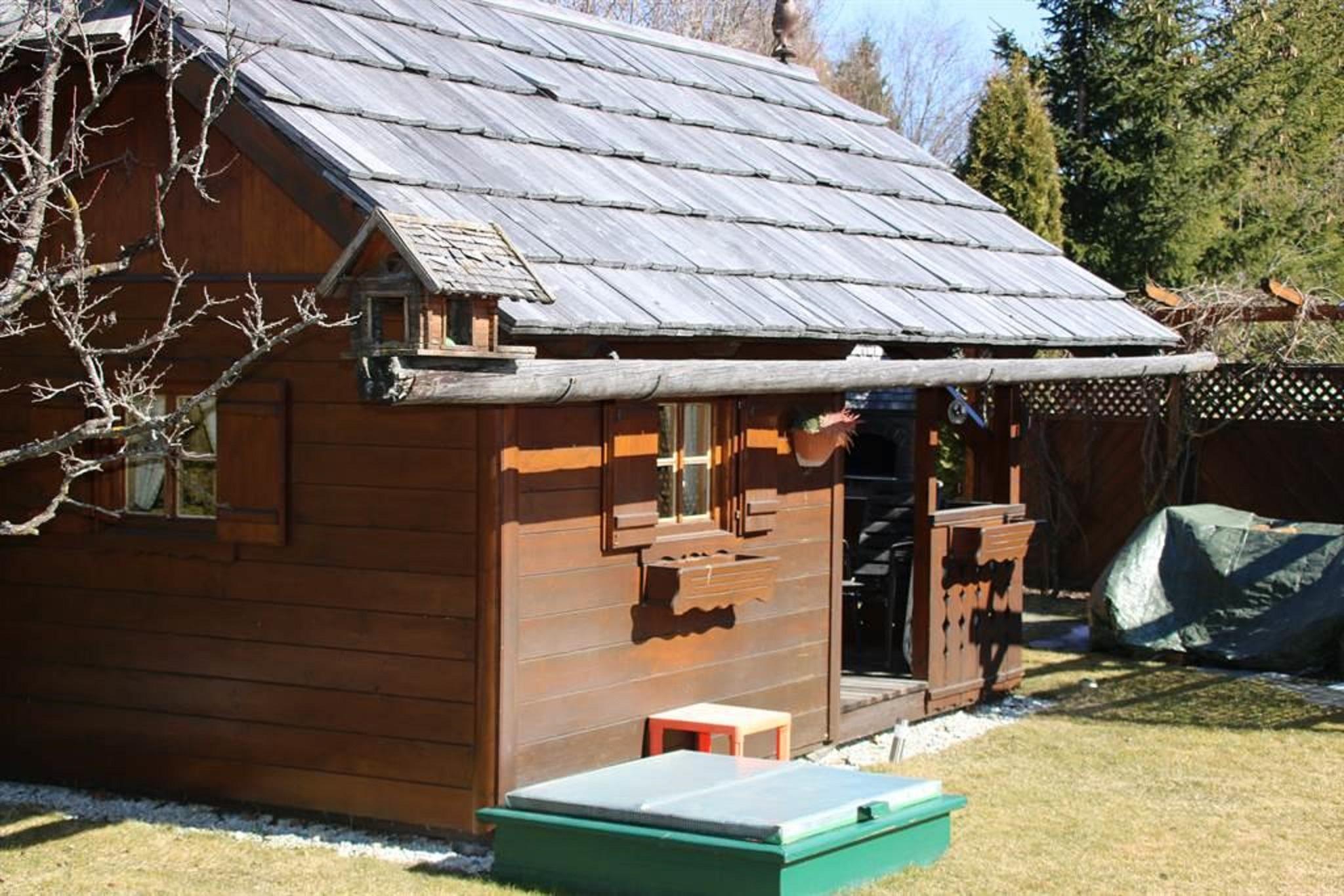 A cozy wooden cabin with a sloped roof and shutters. The garden is well-maintained and there is a green box in the foreground.