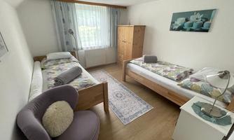 A bright and cozy bedroom with two single beds and a soft seating area. Large windows provide plenty of daylight and the decoration is simple and tasteful.