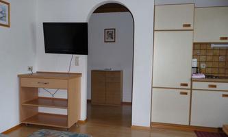 A cozy living space with a TV on the wall and an open access to the kitchen. The furniture is bright and modern, with wooden details.