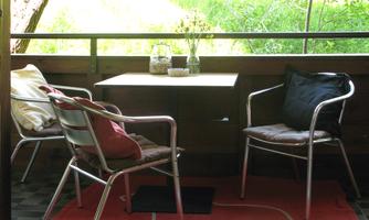 A cozy balcony with two chairs and a table. The floor is covered with a red carpet, and green plants are visible in the background.
