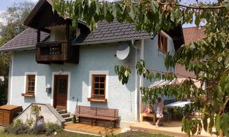A beautiful house with a light blue facade and a balcony. In the foreground, there is a garden with seating and two children playing.