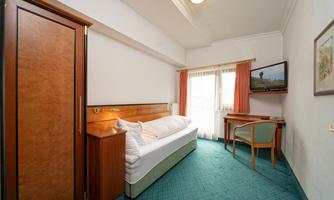 A simple hotel room with a single bed, a table, and a television. The room has large windows with curtains and a green carpet.
