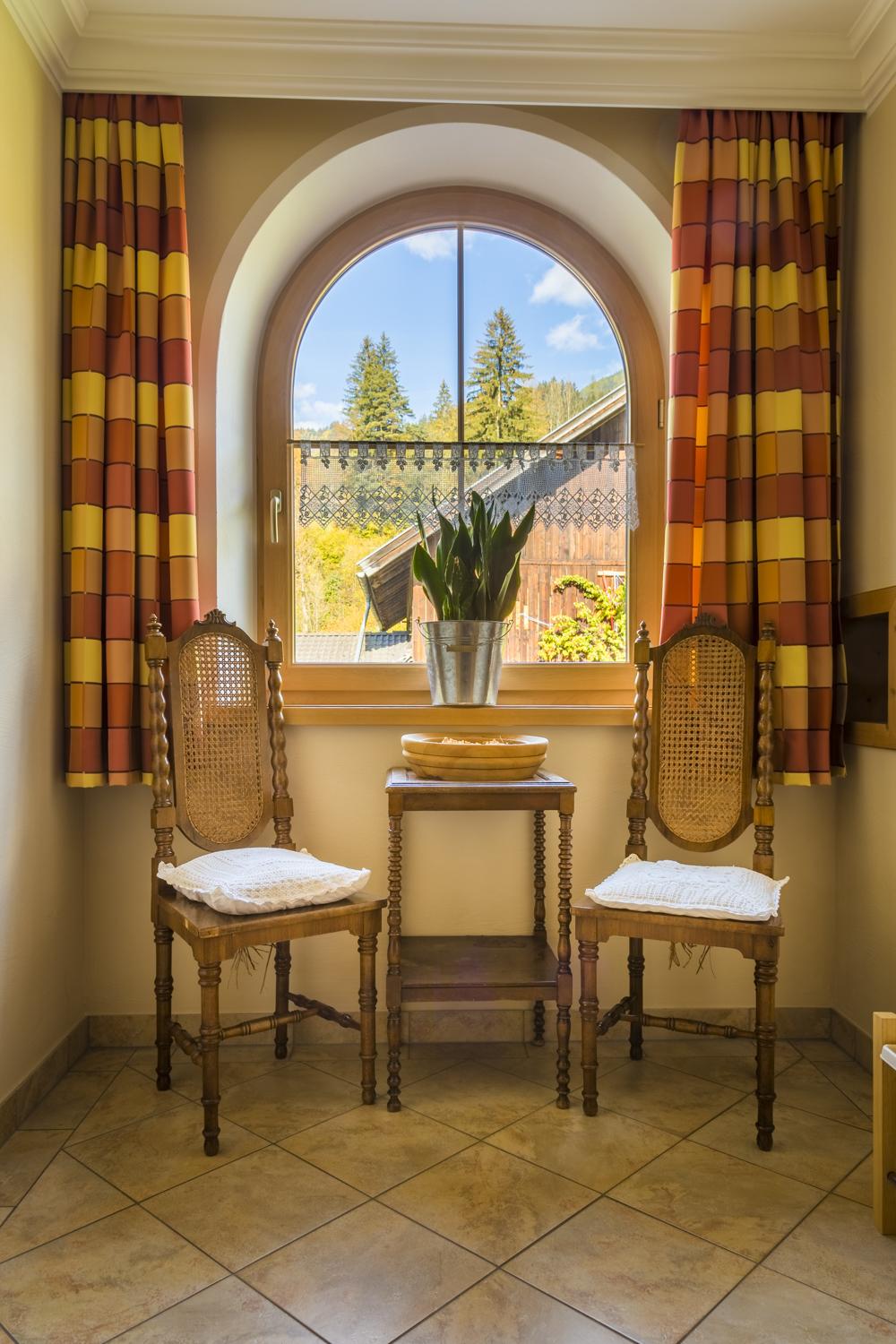 A bright room with two chairs and a small table. Plants are placed in the window, and there are decorative curtains.
