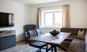 A modern living room with a large dining table and a comfortable couch. A television is mounted on the wall, and a window brings daylight into the room.