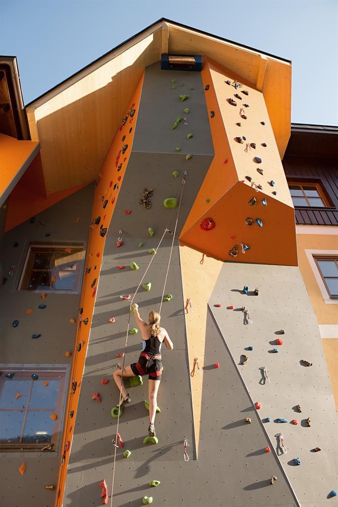 A person is climbing on a colorful climbing wall. The sky is blue, and the building has a modern architecture.