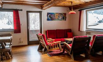A cozy living space with red sofas and a wooden table. Large windows offer a view of a snowy landscape.