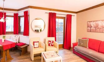 A cozy living room with red curtains and wooden details. There is a table, two armchairs, and a couch that looks inviting.