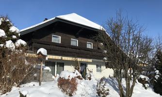 A wooden house in the snow with a blue sky. Surrounded by snow-covered bushes and trees.