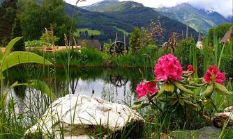 A picturesque pond surrounded by green grass and colorful flowers. In the background, majestic mountains rise under a slightly cloudy sky.