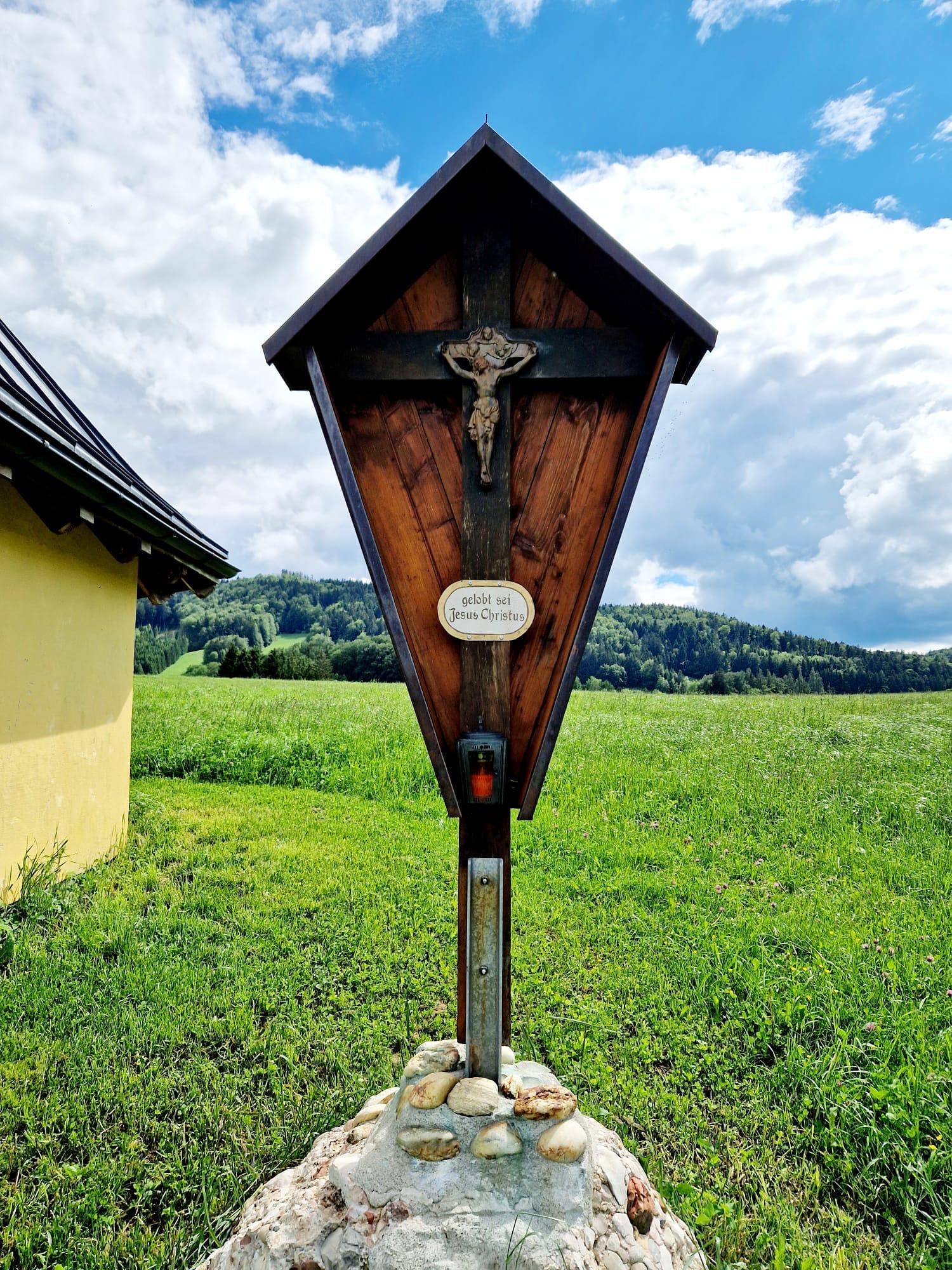 A wooden statue with a cross stands in a green landscape. In the background, gentle hills and a blue sky with clouds can be seen.